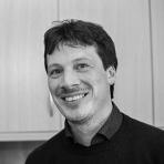 A man with dark hair and a mustache is smiling, standing in front of wooden cabinets.