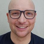 A bald person with light skin, wearing black-rimmed glasses and a black shirt, smiles at the camera against a plain light background.