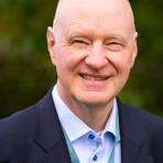 Bald man wearing a dark suit jacket, light blue shirt, and lanyard, smiling outdoors with a blurred green background.