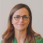 A woman with long brown hair and glasses, wearing a green top, poses in front of a plain light background.