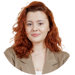 A woman with wavy red hair, wearing a tan blazer over a white top, poses against a plain white background.