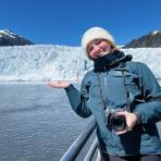 A person in a winter jacket and hat stands on a boat, smiling and gesturing toward a glacier and snowy mountains under a clear blue sky.