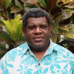 A man with short curly hair wearing a blue and white floral shirt stands in front of large, colorful tropical plants.