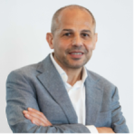 Man in a gray suit jacket and white shirt standing with arms crossed, smiling at the camera against a plain light background.