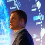 Man wearing a suit and headset microphone speaks in front of a screen displaying satellite images and labels related to Earth observation.