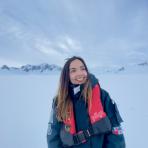 A person in a dark jacket and red life vest stands smiling in a snowy, mountainous landscape under a cloudy sky.