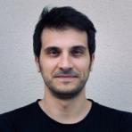 A man with short dark hair and light facial hair is wearing a black shirt and standing in front of a plain, light-colored wall.