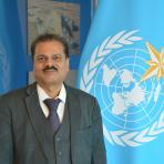 Man in a dark grey suit and patterned tie standing in front of a blue World Meteorological Organization flag.