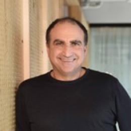 A man in a black shirt smiling in front of a wall.