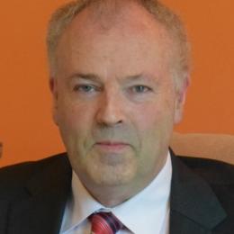 A man in a suit and tie sitting in front of an orange wall.