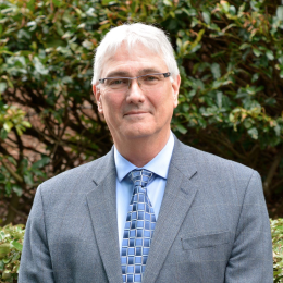 A man in a suit and tie standing in front of bushes.