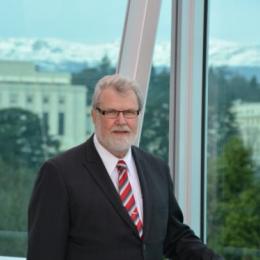 A man in a suit standing in front of a window with mountains in the background.