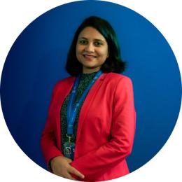 Sulagna Mishra in a red jacket standing in front of a blue background.