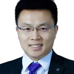 A man with short black hair, wearing glasses, a dark suit, a white shirt, and a purple tie, smiles at the camera against a plain white background.