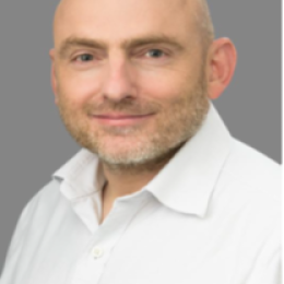 A bald man with a trimmed beard and mustache is wearing a white collared shirt and smiling, posed against a grey background.