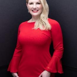 A woman with long blonde hair, in a red dress with bell sleeves, stands against a dark, plain background, smiling at the camera.