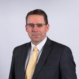 A man in a dark suit, light yellow tie, and glasses poses against a plain white background.