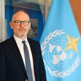 A man in a suit and tie stands next to a flag with the United Nations emblem in an office setting.