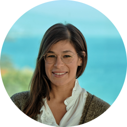 A woman with glasses and long hair wearing a white blouse and gray cardigan stands in front of a blue background.