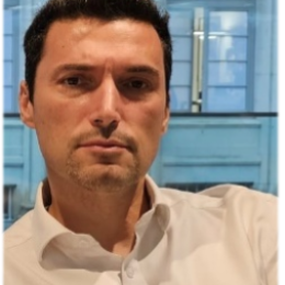 A person wearing a white shirt is looking at the camera with a neutral expression. There is a building with arched windows visible in the background.