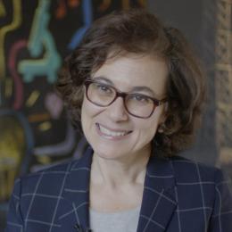 A person with curly hair and glasses smiles, wearing a checkered blazer. A colorful abstract background is visible.