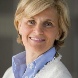 A woman with short blonde hair smiles, wearing a blue collared shirt and a white textured jacket.