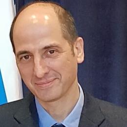A person with short hair is wearing a suit and tie, standing in front of a dark curtain with a hint of a blue and white patterned flag visible.
