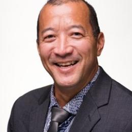 Man in a dark suit and patterned shirt smiling against a plain background.