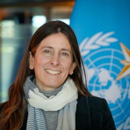 Woman with long hair smiling, standing in front of a blue flag with a global symbol.