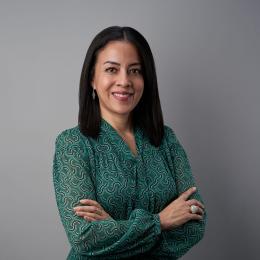 Person with long dark hair in a green patterned blouse stands against a gray background, arms crossed.