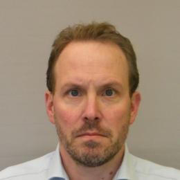 A man with short hair and a beard, wearing a light-colored collared shirt, looks directly at the camera against a plain background.