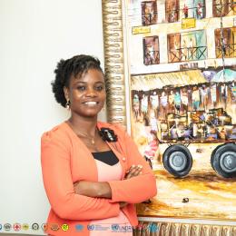 Smiling person in an orange blazer stands with arms crossed in front of a colorful painting depicting a market scene. Various organizational logos are visible at the bottom edge.