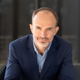 A person in a blue suit and light blue shirt is sitting, gazing at the camera, with a neutral expression against a blurred dark background.