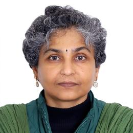 A person with short, curly gray hair and a green jacket looks directly at the camera against a plain white background.