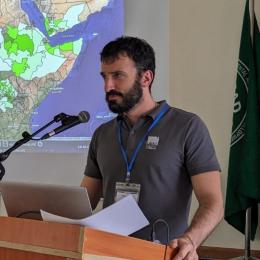 Man speaking at a podium with a microphone, laptop, and papers, in front of a world map projection. Green and white regions highlight areas on the map. Flag partially visible on the right.