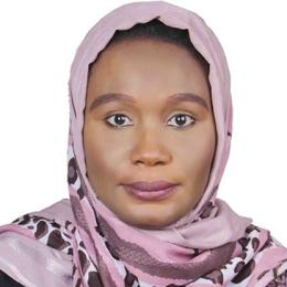 A woman wearing a light pink and brown patterned headscarf, looking directly at the camera with a neutral expression against a white background.