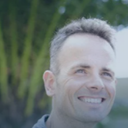 A man with short hair smiling outdoors, with a blurred background of green foliage and blue sky.