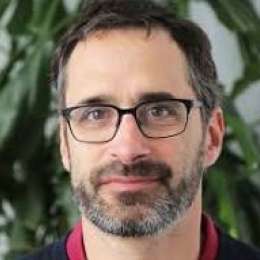 A man with glasses and a beard, wearing a dark top, is in front of a leafy plant backdrop.