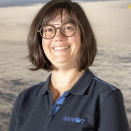 A woman with glasses and short brown hair smiles while wearing a navy polo shirt with the SWOT logo, standing in front of a backdrop showing clouds and space.