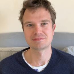A man with short brown hair and blue eyes sits on a gray couch, wearing a navy blue sweater over a white shirt, facing the camera in a well-lit room.