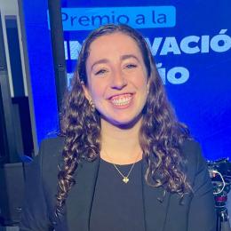 A woman with long curly hair wearing a black blazer and necklace smiles in front of a blue background with partially visible Spanish text about an innovation award.
