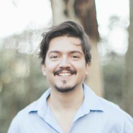 A man with medium-length dark hair and a mustache smiles at the camera outdoors, wearing a light blue collared shirt. Trees and sunlight are visible in the background.