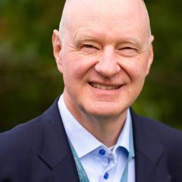Bald man wearing a dark suit jacket, light blue shirt, and lanyard, smiling outdoors with a blurred green background.