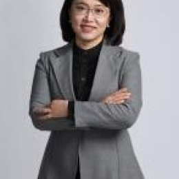 A woman wearing glasses and a gray suit stands with her arms crossed, looking at the camera against a plain background.