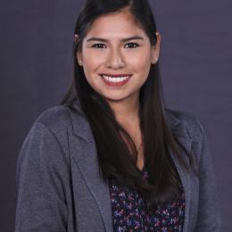 A woman with long dark hair wearing a gray blazer and patterned blouse smiles at the camera against a plain dark background.