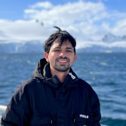A man in a black jacket stands outdoors by a body of water with snowy mountains in the background on a sunny day.