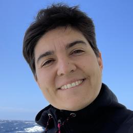 Person smiling outdoors with the ocean and a clear blue sky in the background.
