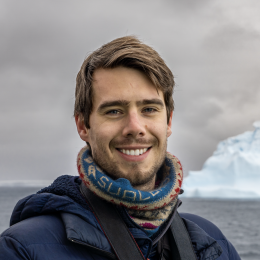 A man with brown hair and a beard, dressed in a blue jacket and multicolored scarf, stands outdoors near icy water with an iceberg in the background.