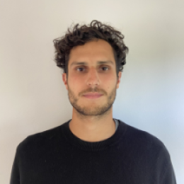 A man with short curly brown hair and a trimmed beard is standing against a plain light-colored wall, wearing a black crew neck sweater.