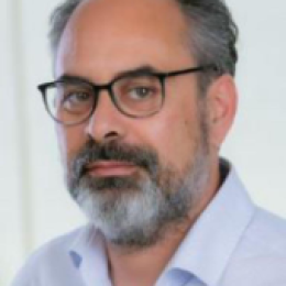 A middle-aged man with glasses, graying hair, and a beard, wearing a light-colored collared shirt, poses in front of a plain background.